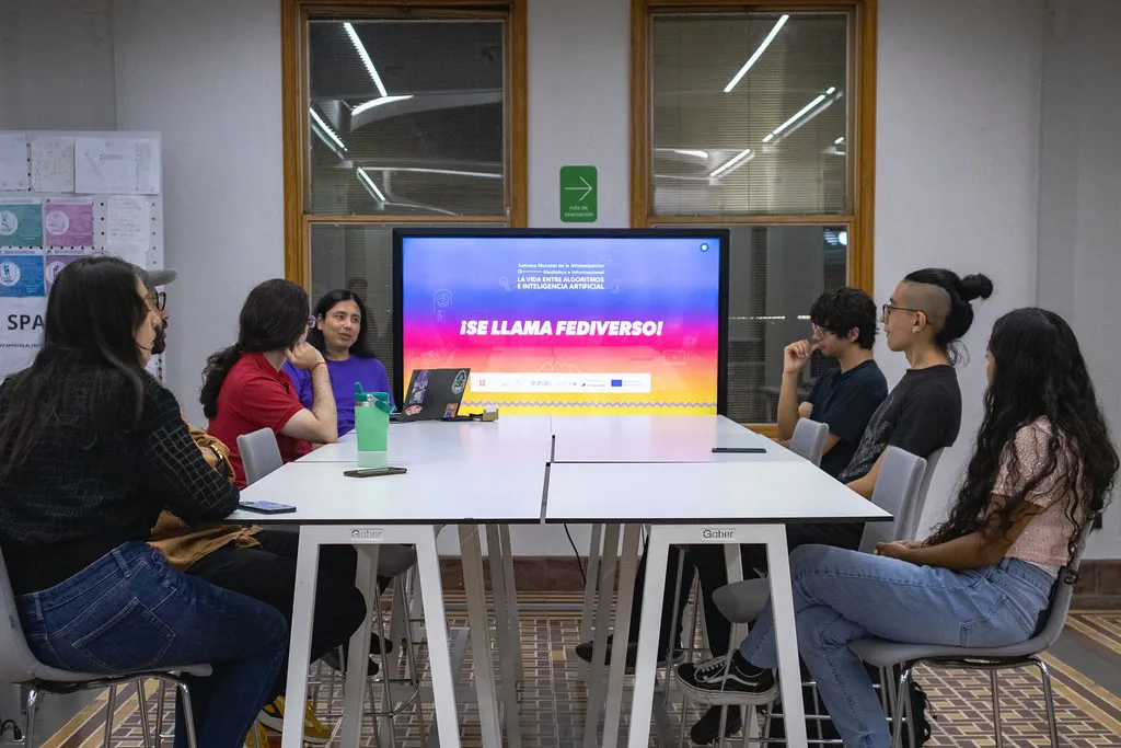 Mesa alta con siete personas sentadas alrededor en el Área de Producción Digital del LabNL, al fondo y al centro hay una pantalla con el nombre de la sesión: ¡Se Llama Fediverso!