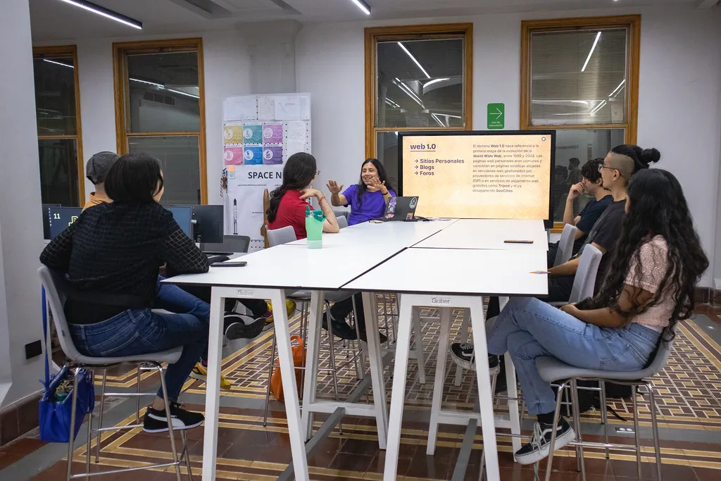 Mesa alta con siete personas sentadas alrededor en el Área de Producción Digital del LabNL, al fondo y al centro hay una pantalla con el nombre de la sesión: ¡Se Llama Fediverso!