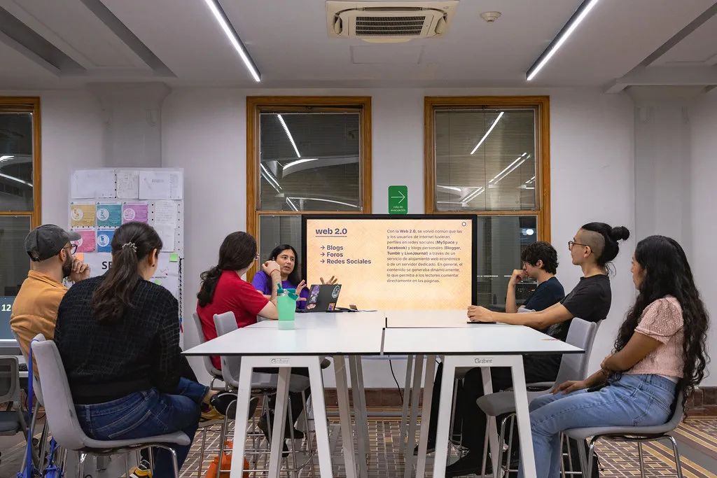 Mesa alta con siete personas sentadas alrededor en el Área de Producción Digital del LabNL, al fondo y al centro hay una pantalla con un texto breve sobre la Web 2.0