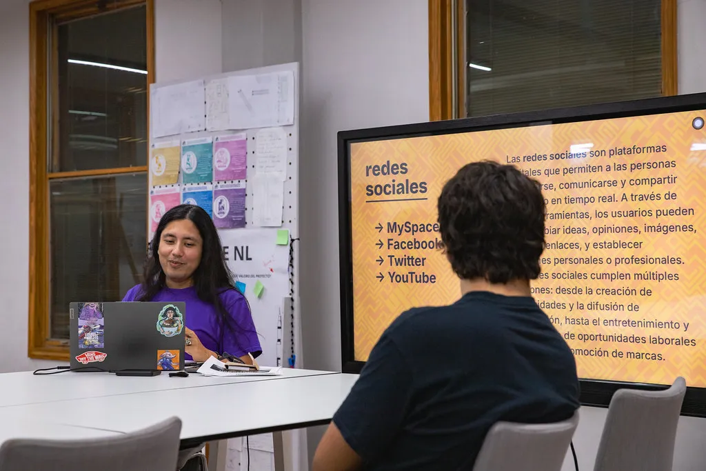Plano medio de una mesa alta con dos personas sentadas alrededor en el Área de Producción Digital del LabNL, al fondo (del lado derecho) hay una pantalla con un texto esplicando las primeras redes sociales.