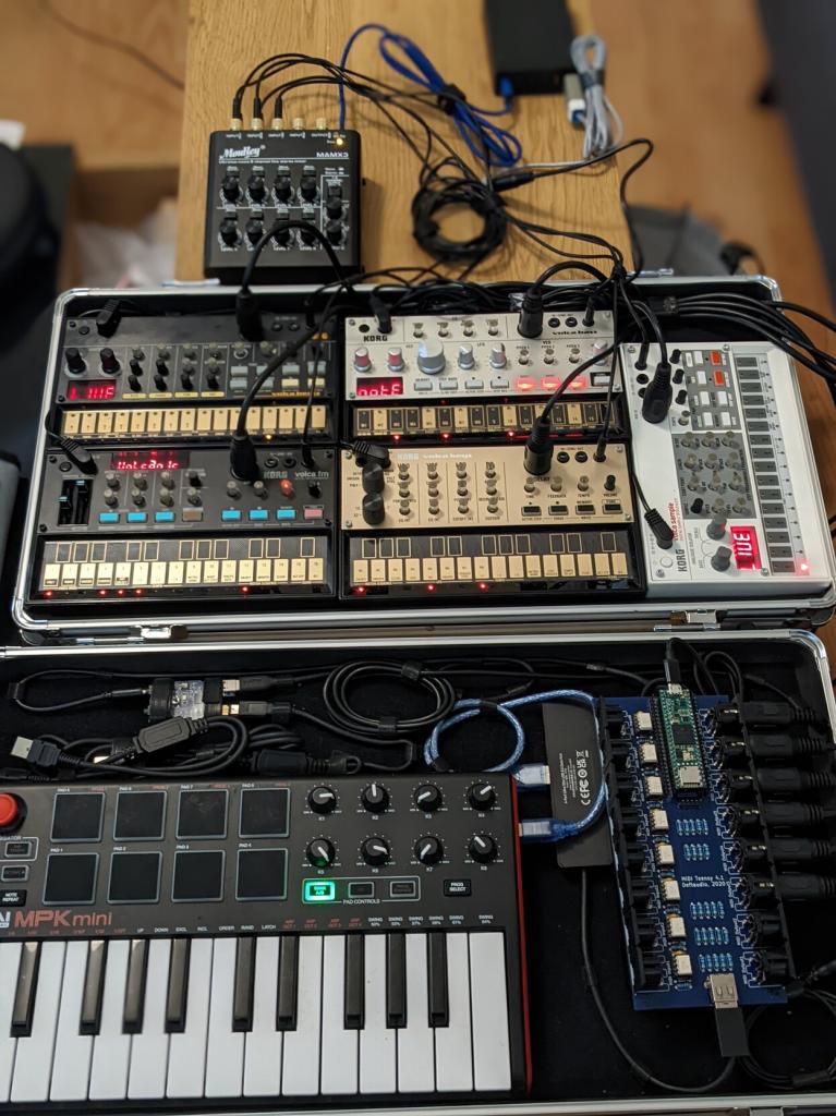 Two gear boxes which have had the tops taken off to reveal 5 Volcas, a MPK mini, an 8x8 MIDI PCB, and other miscellaneous devices and cables, all kept tidy with velcro.