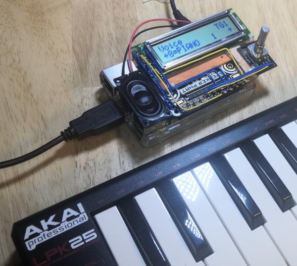 A Pimoroni Speaker Phat mounted on a Raspberry Pi 2. It has been extended with a 1602 liquid crystal character display and a rotary encoder knob. Blue and yellow wires have been soldered to the shared 40-pin heart of the Phat to connect the display and encoder to the Raspberry Pi. Red and black wire emerge from under the extended Phat to a small membrane speaker. An Akai LPK25 MIDI keyboard is plugged into one of the USB ports. The display reads: "Voice TG1 <8=PIANO 1>"