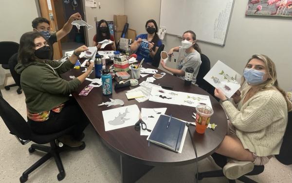 Several lab members sitting around a conference room table cutting out marine creatures (urchins, hammerhead sharks, sting rays, fish, dolphins) and holding them up for the camera 