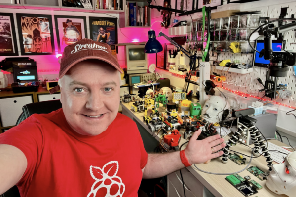 Kevin wearing a red Raspberry Pi shirt and a hat that says Streamer on it. Sat at a desk in his workshop gesturing to a table full of robots and live streaming equipment