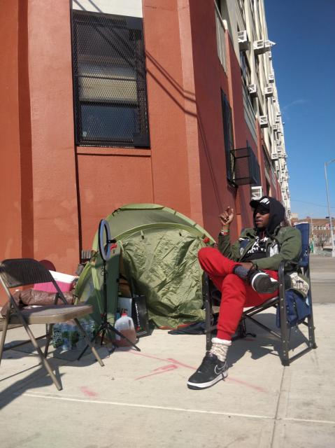 Shuga ray takes a moment to talk to me about the trends in development he's seeing in our neighbourhood. He's wearing red pants and is sitting across from an empty chair, where I was sitting.  Behind us there is a tent and the underutilized DOE building. We had a good time connecting and thinking about how we can support a struggle for justice with art and other collaborative acts.