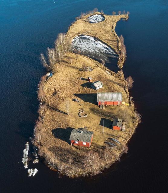 Small, teardrop shaped island with five buildings of various sizes (possibly a house with barns and outbuildings) on the lower right side. The point of the drop has a little round pond, then a very small, nearly enclosed inlet. There are trees on the left, below the closed side of the inlet and a series of shrubs around the edges of the island. Spring is browned grass and leafless trees and shrubs. There is still a bit of ice on the surface of the pond and inlet.