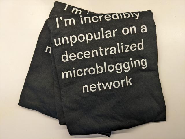 Two black shirts, folded, one sitting on the other at an angle.  In the front is white sans serif text that reads:

"I'm incredibly
unpopular on a
decentralized
microblogging
network"