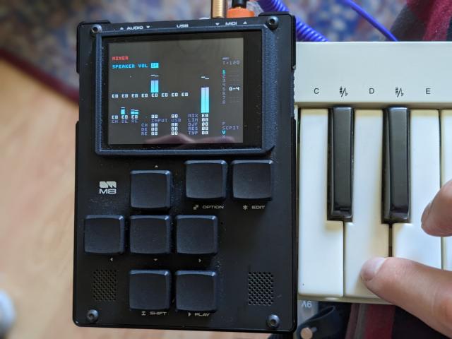 An M8 sitting on top of a Commodore MK-10 keyboard, a purple cable goes from the keyboard to the M8 MIDI in port. A key on the keyboard is being pressed and the M8 mixer shows that audio is being played.