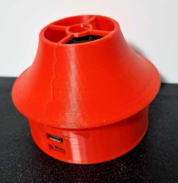 A small palm sized red sensor module sits on a desk. The red plastic case is slightly rough indicating it has been 3d printed. Visible on its sides are 2 access ports for usb cables - for charging and connecting to a computer. Visible on the modules conical top is a small opening with a fan enclosed. This draws air into the unit and pushes it over the sensors.