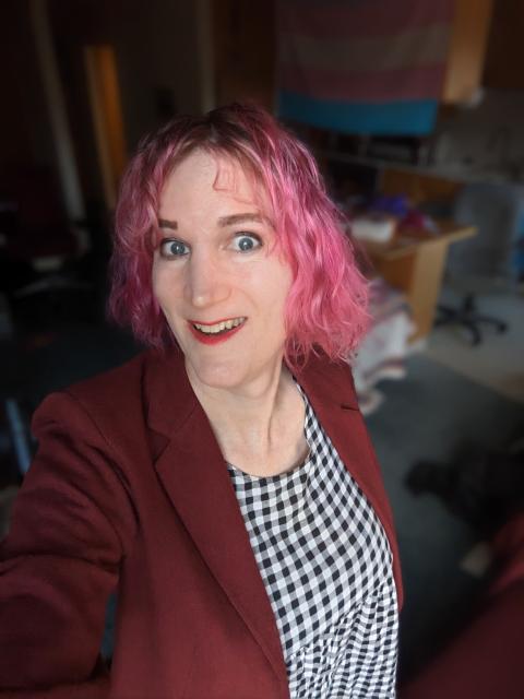 A portrait of Charlie Jane Anders wearing a red jacket and check dress in front of the trans flag