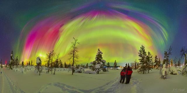 Two people dressed in red coats are standing on a snowy landscape with bare trees. Above, many aurorae of different colors appear, with some stars visible in the background.