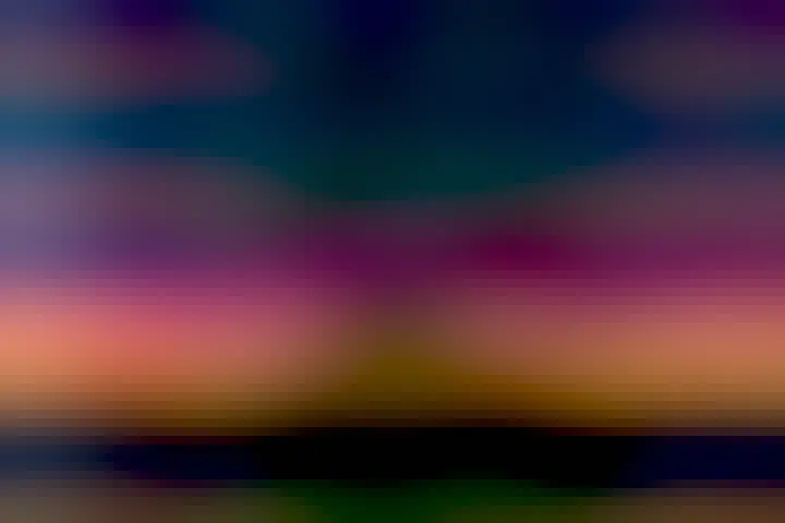 A dark rectangular building is seen across a green field with colorful aurora, mostly red, seen in the background sky. Past the aurora, the sky is also filled with stars.