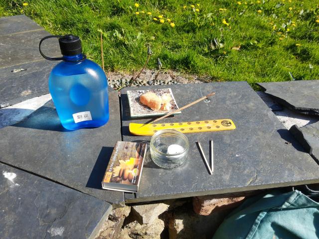 Stone seat with a little glass candle holder, an incense stick, a small mosiac covered plate with some cake on, and a large blue water bottle on the left, there is green grass and dandelions in the background.