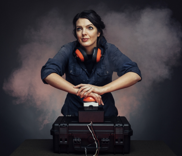 Fran pressing a big red button with smoke from an explosion wisping around her. She is wearing a navy blue boiler suit and has big orange ear defenders round her neck.