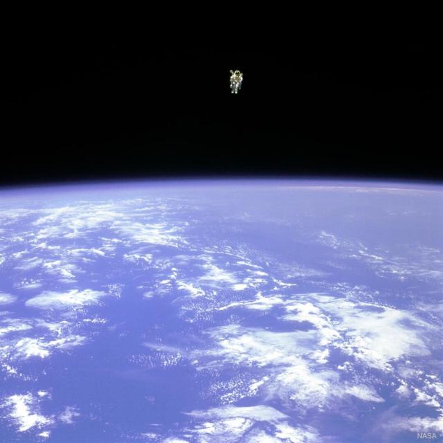 An astronaut is seen hovering over the Earth. In the top part of the image, the astronaut is seen against the darkness of space. In the lower part of the image, the Earth is bright blue with white clouds.