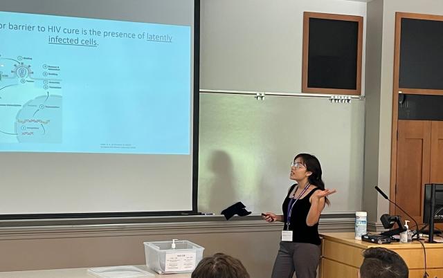 A person near a podium looks at a screen that says "the major barrier to HIV cure is the presence of latently infected cells" as she speaks to an audience in a classroom