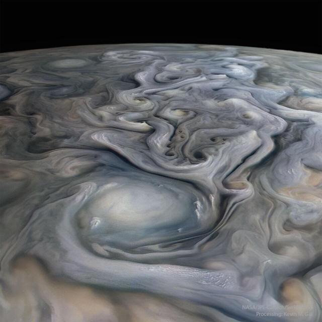 The cloud tops of Jupiter are pictured in a closeup flyby of the Juno spacecraft. A big white oval cloud is visible in the foreground, while many swirls of many muted colors are visible trailing behind. A dark night sky is in the background.