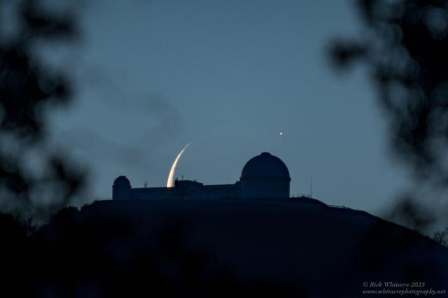 A dark mountain lies in the center with an observatory building sporting two telescope domes. The background sky appears dark blue. Behind the center of the observatory is part of a crescent moon, with an unusual bright spot to its upper left.