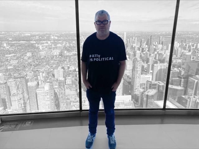 Me, standing on the viewing floor at top of CN Tower in Toronto. I am wearing my #a11y is political t-shirt. Behind me, a view of Toronto.