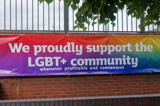 Photo of a banner with a rainbow background and white text which says "We proudly support the LGBT+ plus community" and underneath in smaller letters "whenever profitable and convenient"