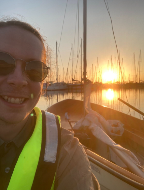 a happy cblgh in a neon yellow floatation vest. in the background the sun is setting. the boat with the main sail having been lowered is present, with one of the oars in frame