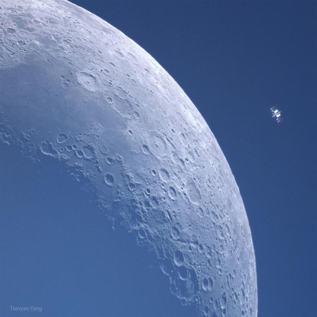 A crescent moon is shown against blue background. Many craters are visible in great detail. To the upper left appears some kind of small machine which is actually the International Space Station also in orbit around the Earth.