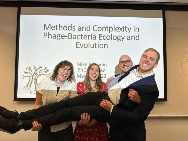 My three siblings holding me up in front of the title slide of my defense: "Methods and Complexity in Phage-Bacteria Ecology and Evolution"