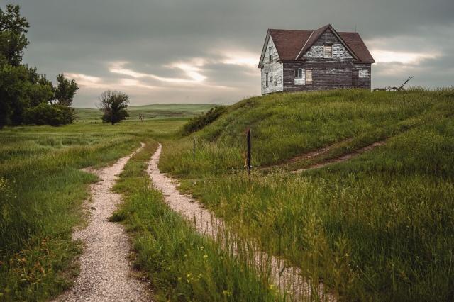 An old farmhouse with weathered paint sits on a hill overlooking a road and a beautiful skyline.