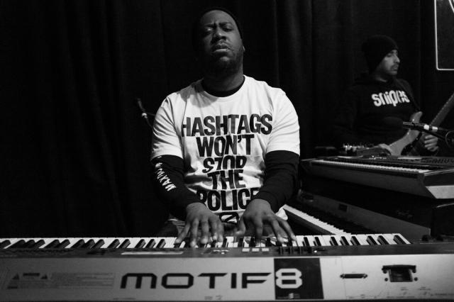 A Black male pianist (Robert Glasper) facing the viewer, with eyes closed, a toothpick in his mouth, and both hands playing a keyboard. He is wearing a t-shirt reading "Hashtags Won't Stop The Police" in all caps.