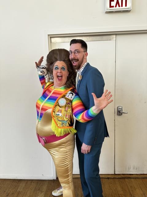 A tall bearded man in a teal suit excitedly poses with international lady rapper superstar Leslie Hall who is making jazz hands while wearing gold spandex pants and a rainbow spandex top