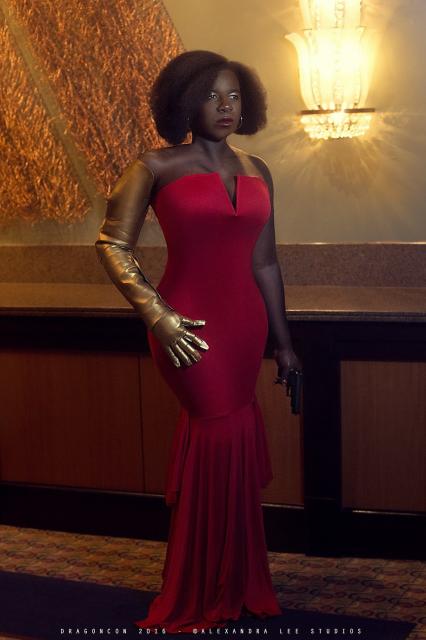 photo of a dark-skinned Black woman (Misty Knight) with an afro, wearing a strapless red floor length mermaid style wiggle dress, and a gold metal right arm, while holding a small pistol in her left, natural arm. There are gold plated panels on the wall and a bright light fixture behind her. Text on bottom reads "DragonCon 2016  © Alexandra Lee Studios"