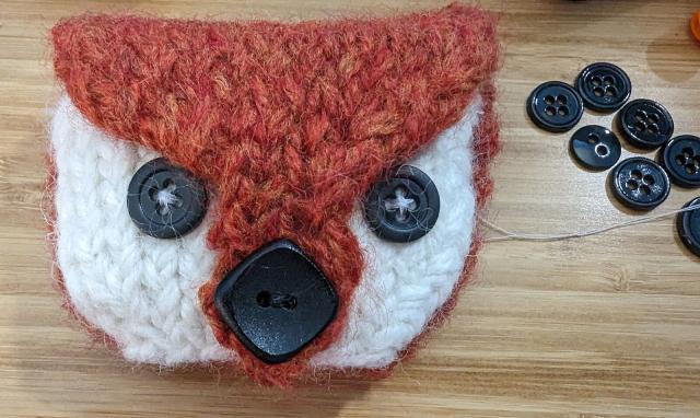 A felted wool pouch in the shape of a fox's head with round black buttons for eyes and a larger diamond-shaped black button for a nose (which is the clasp of the pouch). To the right are a small collection of round black buttons and some white thread.