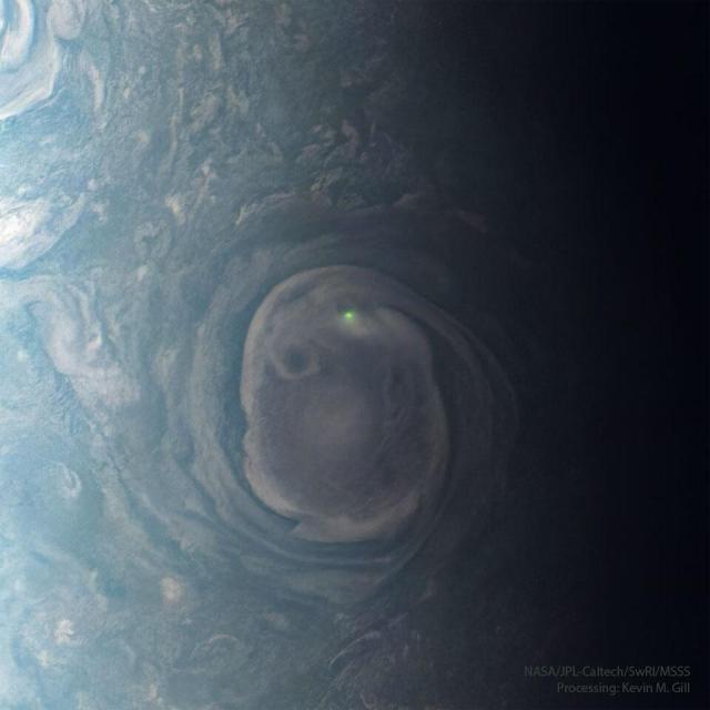 A large swirling cloud on Jupiter is shown with a bright green spot near its top. The cloud is surrounded by other less descript parts of Jupiter's upper atmosphere.
