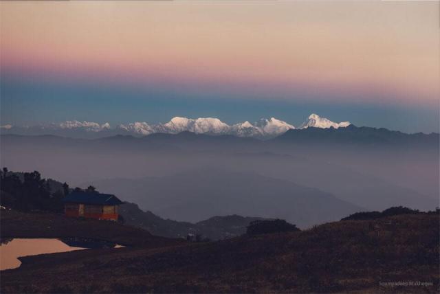 An orange sky hovers above snow-covered mountains. A blurry line divides the orange sky from a darker sky. In the foreground are hills and a house.