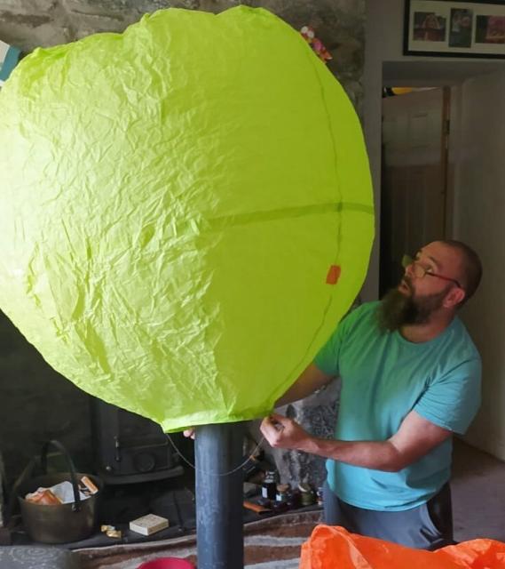Bearded man kneeling in an old N Wales cottage, he has a chimney contraption in front of him and a large green tissue paper hot air balloon filling with hot air!