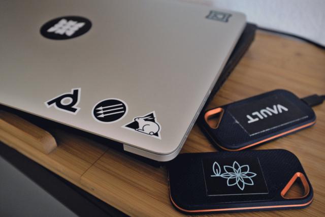 A 2012 Macbook and two small external harddrives on a wooden surface. Both have some of the stickers of the previous image applied to them.