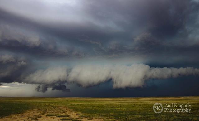 A shelf cloud rolls over the plains. The cloud is grey and back-lit in shades of blue and green. The fields are treeless and shades of green to gold.