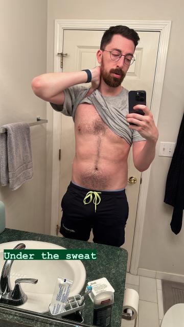 A tall white bearded man in front of a mirror wearing athletic gear, lifting his shirt to show a sweaty torso with the caption “under the sweat”