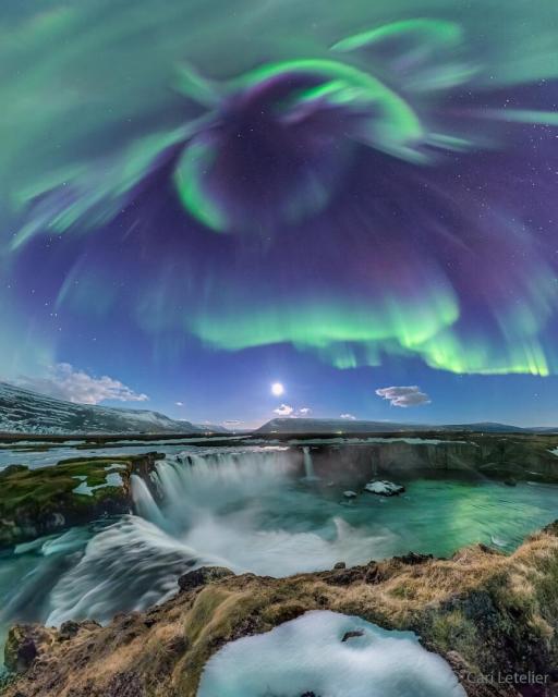 Green aurora are seen against a blue sky over a series of picturesque waterfalls.