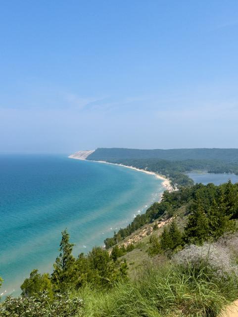A view of Lake Michigan and the coastline from an elevated point on a park trail. The water is dark far from shore but grows progressively lighter in the shallow water. The coast is mostly forested, except for the beaches and a prominent, steep dune in the distance.