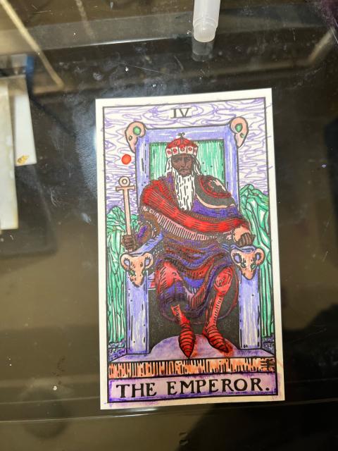 Colored tarot card. A man in a crown sits on a throne. He wears draped reds, preaches, and blue-purples before a green and purple landscape. He is Black with white long hair and beard, he seems quite fashionable IMO. The bright landscape is meant to be joyous and compliment him, not be oppressed or boring under his rule.