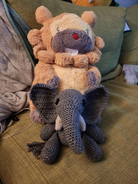 A grey elephant amigurumi sitting in front of a bidoof plush on a couch.