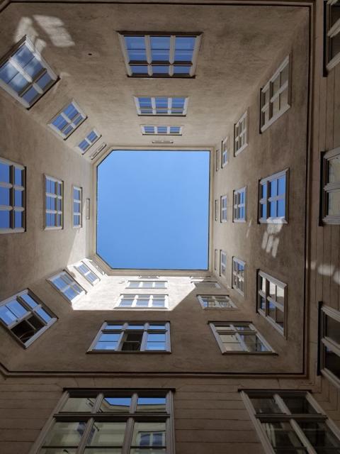 Fotografie eines engen Innenhofes von unten kerzengerade nach oben. Der Himmel ist in der Mitte des Bildes strahlendblau, viele Fenster DES Gebäudes reihum.