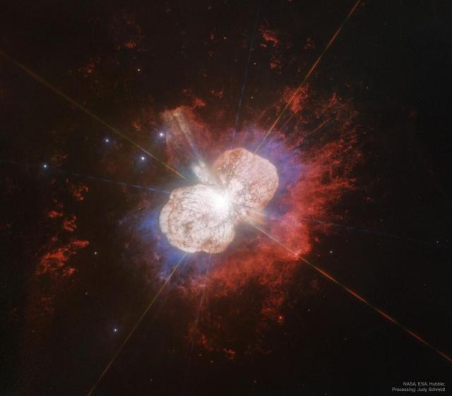 A Hubble image of the gas and dust surrounding the star Eta Carinae is shown. The nebula has two distinct light-colored lobes, surrounded by red glowing gas.