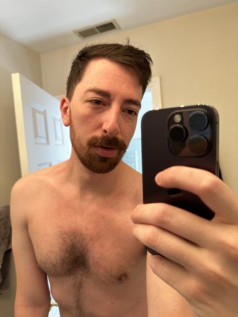 Jonoor poses in front of a bathroom mirror shirtless with a thick mustache and shorter beard facial hair.