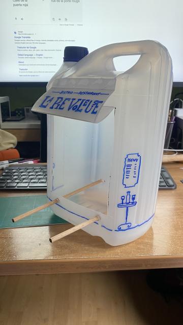 Photo of a bird feeder made from a recycling five litre jug. On side has been cut to make the entrance. The discarded plastic has been used to make awning. It has two wood beams for the bird to land. It is lightly decorated as a French bistró with marker drawing of a table with a bottle of wine and two cups. On the awning it reads “Bistró Restaurant La Pie Voleuse” (the thieving magpie)