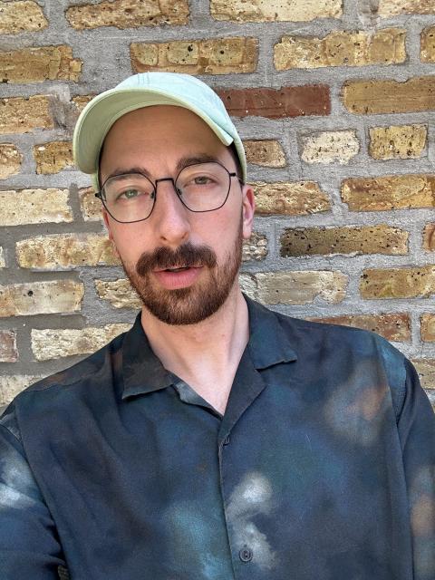 A bearded man with glasses wearing a mint green hat and dark blue patterned button down shirt poses in front of a brick wall