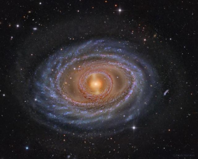 A spiral galaxy is shown with a yellow center, blue rings and spiral arms, and dark brown and red dust. The surrounding dark field contains both local stars and more distant galaxies.