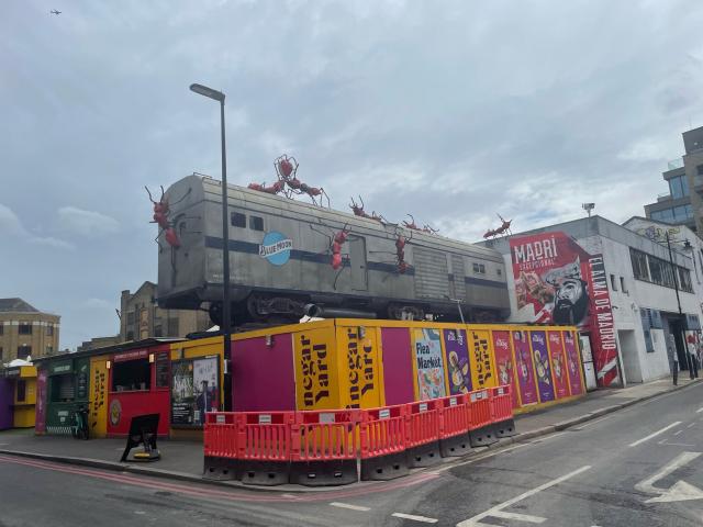 How do I describe this, well it’s a train car on top of a street restaurant building covered in giant red ant sculptures that look like they’re climbing all over the train there is one on top of the building behind the train and a pair at the front who are facing each other and holding a red heart over their heads with their jaws