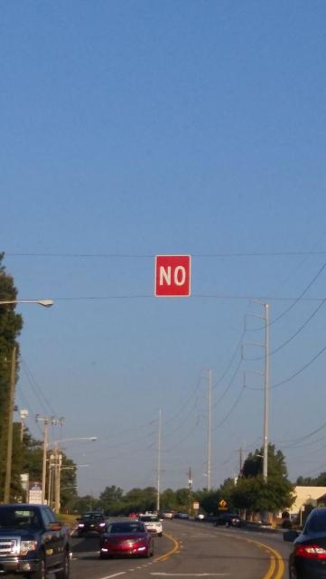 A two-way suburban road with cars on it. Suspended above and, for no apparent reason, is the single word "NO" - in white letters on red indicating a traffic rule. The pic is very vertical, so the sign is framed against an expanse of clear blue sky
